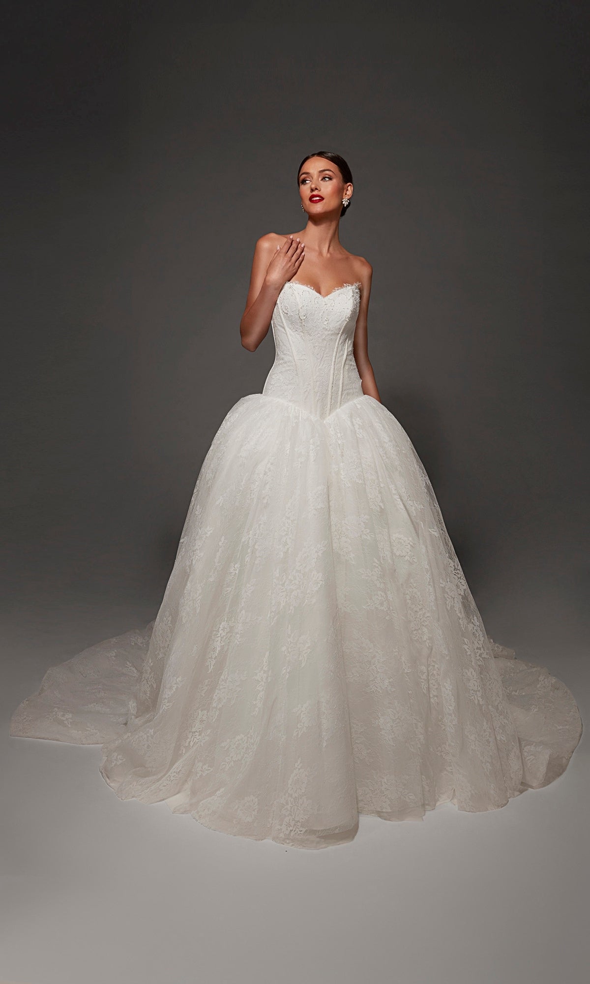 A woman poses against a dark background wearing the Long Wedding Dress: Alyce 7169, a strapless lace ball gown featuring a fitted bodice, full lace skirt, romantic sweetheart neckline, and a long train.
